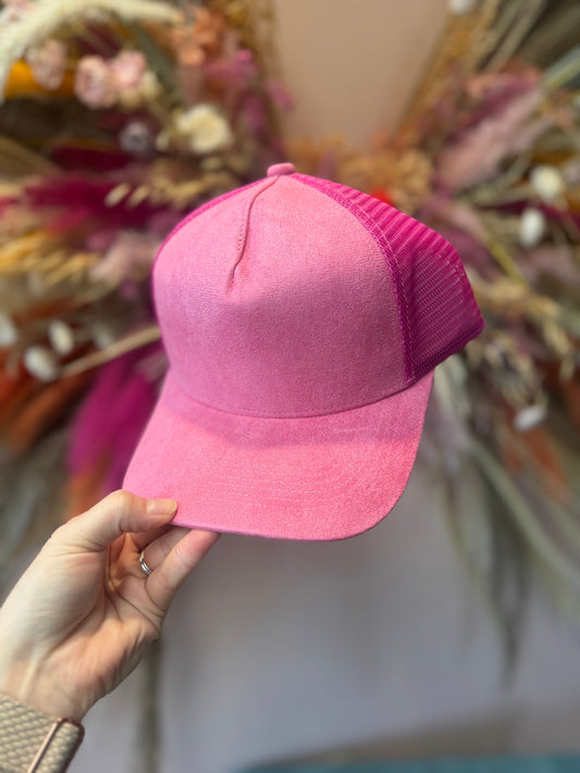 A hand holding a hot pink trucker hat with vegan suede front and mesh back, displayed against a floral background.