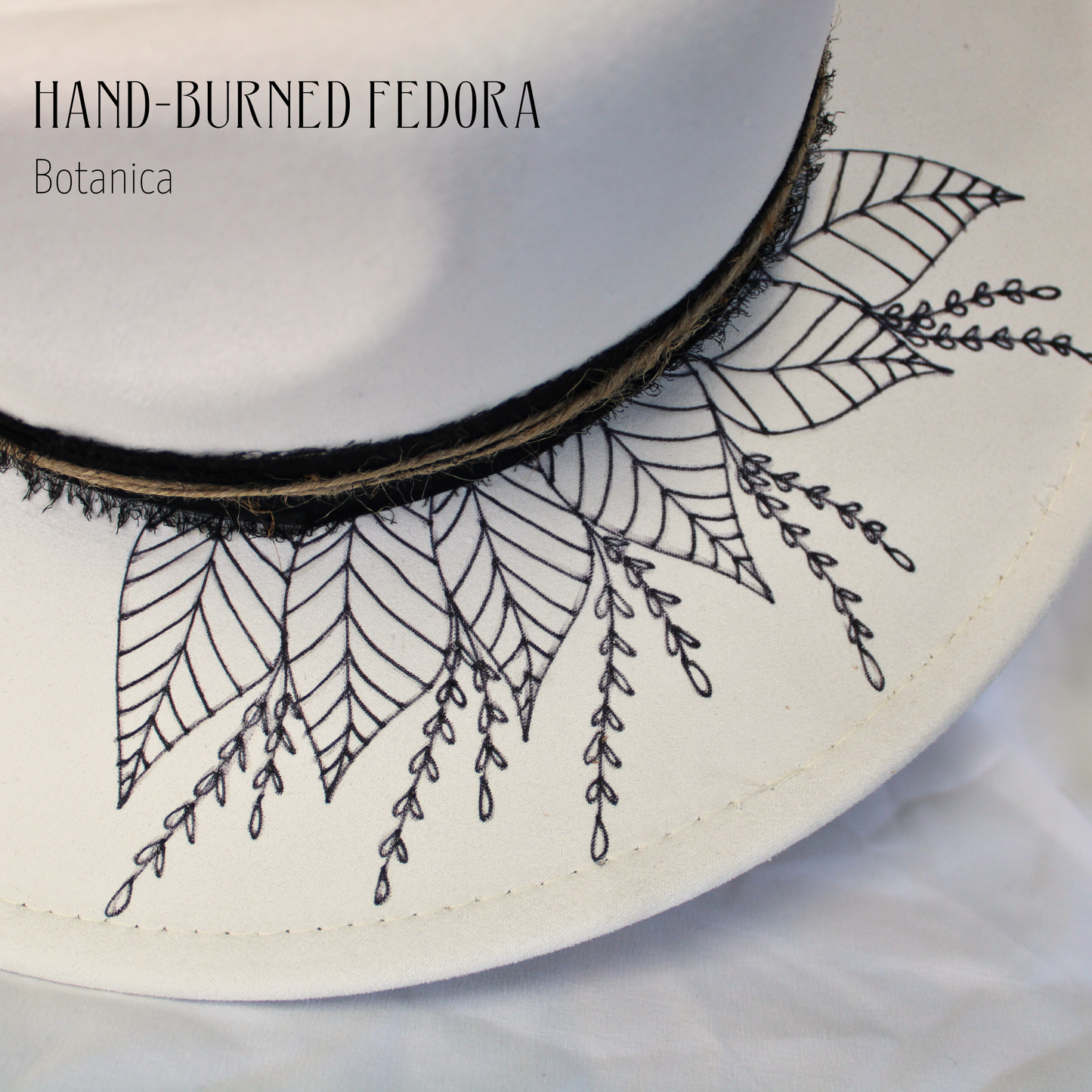 White hat with black embroidered leaf patterns on a white background, featuring 'Hand-Burned Fedora Botanica' text.