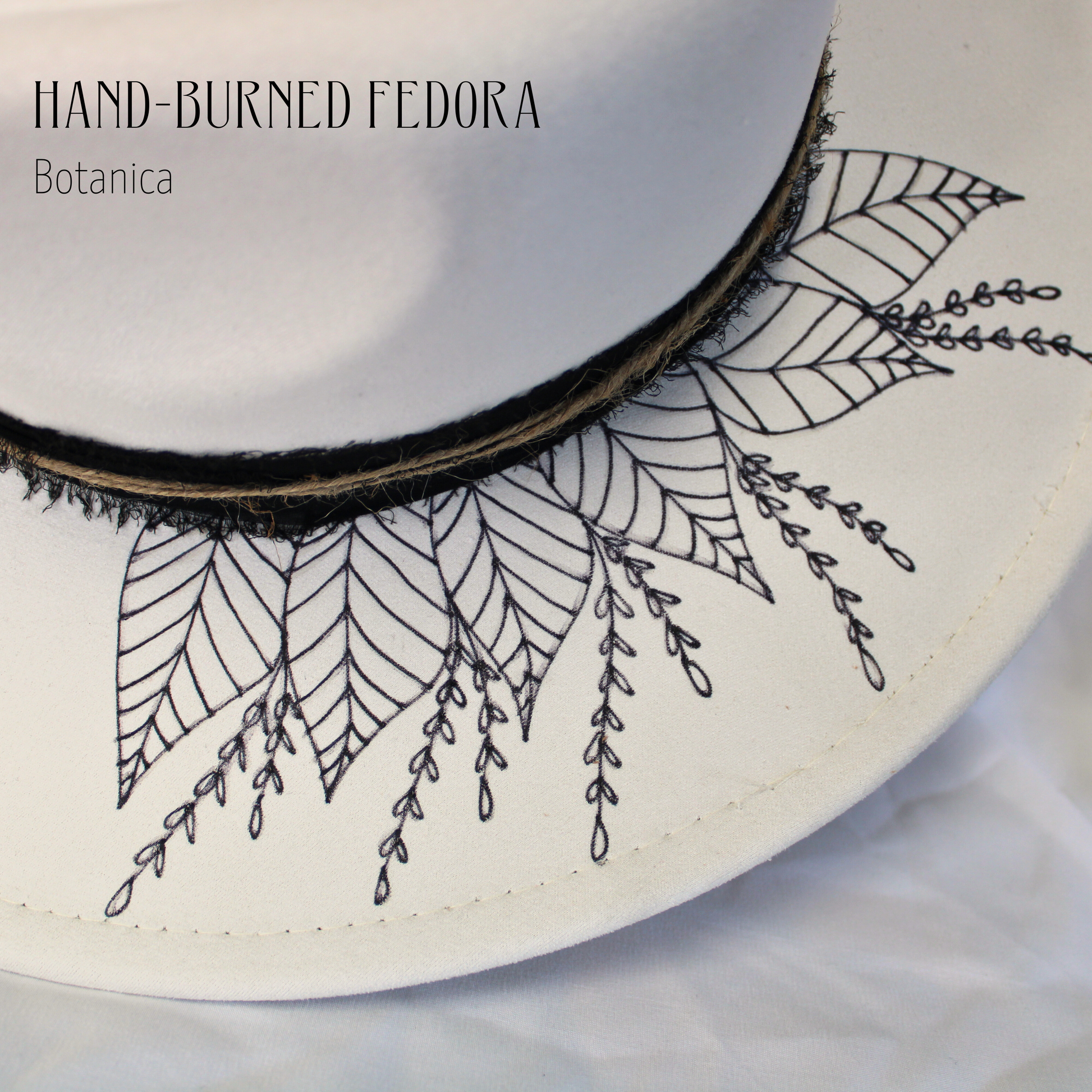 White hat with black embroidered leaf patterns on a white background, featuring 'Hand-Burned Fedora Botanica' text.