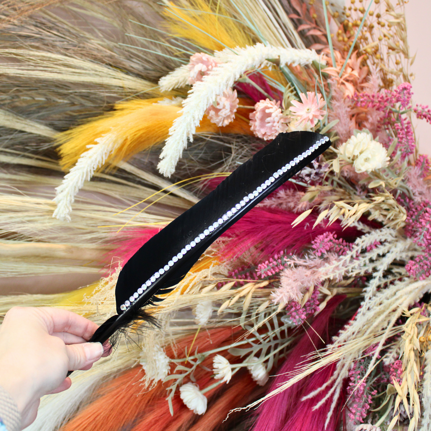 A black turkey feather adorned with rhinestones being held by a hand against a backdrop of various dried flowers and feathers.